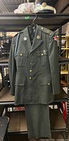 Full view of the vintage US Army dress uniform jacket with scarf and matching pants on hanger against storage background.