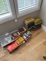 Stacks of assorted books piled on a hardwood floor. Various titles and sizes visible, showing a mix of hardcovers and paperbacks, some stacked flat others upright.
