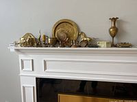 Overall assortment of brass decor items shown on a white mantel against a wall.