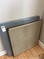 Two folded card tables stacked against a wall, showing different surface materials: one beige fabric, one gray plastic.
