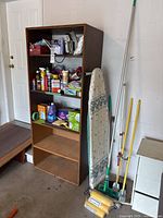 Wooden shelving unit filled with assorted cleaning supplies and placed next to ironing board and cleaning sticks used for cleaning purposes