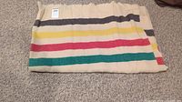 Folded view of the cream woollen blanket showing green, red, yellow, and black stripes along one edge and a Hudson Bay tag.