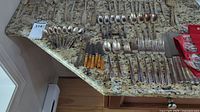 Assorted cutlery laid out on granite countertop including spoons, forks, knives with orange plastic handles, and serving utensils.