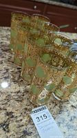 Eight decorative gold and frosted green tumbler glasses arranged on a granite countertop with a MaxSold label nearby.