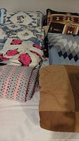 Photo showing various quilts, crocheted blankets and a solid color comforter folded and stacked on a bed, highlighting different textures and patterns.