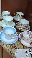 Set of six fine china teacups and saucers arranged on granite countertop showing varied floral and gold accent designs.