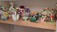 Assorted ceramic floral arrangements and figurines with pastel colors on wooden shelf