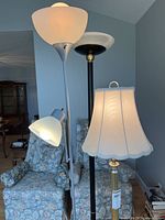 Photo showing three floor lamps placed behind a armchair, includes a brass lamp with a large white scalloped shade, a black torchiere lamp, and a white lamp with a smaller adjustable lamp attached.