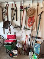 Wall-mounted gardening tools including pitchfork, shovels, rakes, pole pruners, bird feeder, and Scott’s seed spreader on floor.