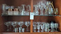 Wide view of cabinet shelf showing various clear and cut glassware: tumblers, stemmed wine and cocktail glasses, shot glasses