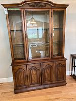 Front view of the wooden hutch showing three glass doors on top, wooden four door cabinet on bottom, ornate top crown molding.