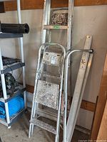 Photo shows two aluminum stepladders of different heights and Little Giant Ladder Wingspan accessory leaning against a wall with other tools visible nearby.
