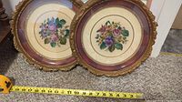 Two of the three round framed Peti Point floral embroidery artworks side by side on carpet with measuring tape showing around 12 inch diameter