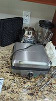 Waffle maker with waffle plates, hand mixer, and blender shown on kitchen counter.
