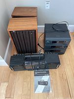 Photo showing RCA music system stack components with control buttons and cassette tray, along with two wooden Realistic speakers and a Sharp cassette radio recorder on floor with manual.