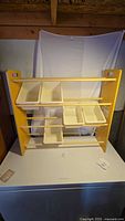Front view of yellow wooden bin storage stand with 5 white removable plastic bins placed on three dowels