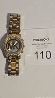 Front of gold tone Michael Kors watch showing the MK logo on the face next to maxsold number card.