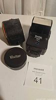 Vivitar 2850 camera flash, lens cap with logo, and black leather carrying case for the lens.