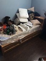 Full side view of the bed frame showing mattress wrapped in plastic with plush toys and pillows on top. Visible are three storage drawers underneath the mattress.