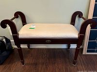 Full view of wood framed bench with cream upholstered cushion, curved armrests, and spindle legs on wooden floor.