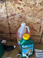 Two chemical containers on a surface with a rough wood panel backdrop; one labeled Weed B Gon and another translucent jug with orange liquid and white cap