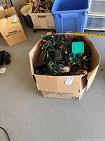 Photo showing full cardboard box containing bundled strands of multi-color Christmas lights, multiple black electronic timers, a green timer box, and cords.
