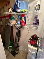 Photo of corner storage with wire shelves holding assorted cleaning chemicals and mop, vacuums on floor, and partially visible ironing board.