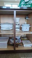 Overall view of the cabinet shelf showing two Rival crock pots, Proctor Silex coffee maker, folded table linens, plastic container, and cookbook.
