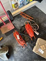 Two extension cords on black rubber floor near garage items, one orange heavy duty cord coiled on orange reel holder, white cord beside it.
