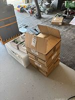 Stack of four cardboard boxes of ceiling lights in a garage setting with other garden tools and equipment visible.