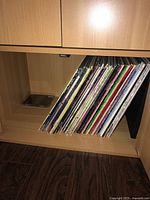 Side view of a wooden shelf with approximately 30 vinyl LP record albums stacked vertically, showing varied colorful spines.