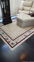 Photo of the full area rug on hardwood floor beneath a beige sectional sofa and recliner, showcasing the floral border design and red striping.