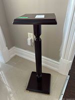 Full view of the wooden stand showing the square top, fluted central column, and square base on a tiled floor with wall in the background.