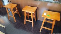 Three wooden stools placed on dark hardwood floor next to a granite countertop. Stools have a rectangular wooden seat and four legs with horizontal support braces.
