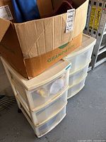 Two white plastic drawer units, each with three drawers and a cardboard box labeled 'GRAND & TOY' on top, containing personal items inside drawers and box.