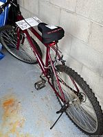 Rear and side view of the maroon men's Supercycle bicycle showing the CCM Mega Gel saddle and the knobby tires. Bike kickstand down, parking against concrete wall.