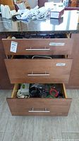Three drawers of kitchen items. Top drawer with small kitchen appliances like Magic Bullet blender and hand mixer cords visible. Middle drawer with cookbooks and kitchen items. Bottom drawer with glassware, small containers and kitchen accessories.