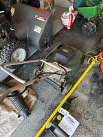 Photo of the green and black Yardworks electric lawn mower placed on a garage floor among other lawn and garden equipment.