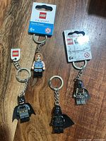Four LEGO character minifigure keychains on wood surface; Rachel Green and The Mandalorian on packaging cards, Batman and Darth Vader loose