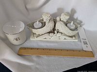 White scented candle in glass with AB logo, silver lid beside angel candle holder and ruler.