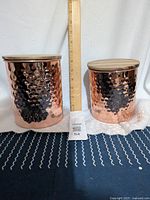 Front view of two hammered copper containers of different sizes with wooden lids placed beside a ruler for size reference.