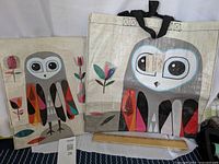Two reusable bags with owl designs made of woven recycled plastic material - one grocery tote bag and one mail bag style with shoulder strap; items are flat and handle details visible.