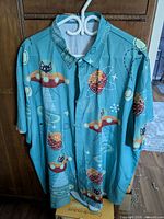 Front view of turquoise short sleeve button-up shirt with black cartoon cats inside orange UFOs and atomic orbit patterns