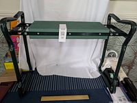 Front view showing stool set up flat with larger green cushion on top as seat, black steel frame and handles on sides.