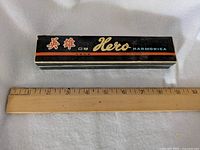 Closed black original box of Hero harmonica with Chinese text and colored stripes, alongside 12 inch ruler for scale.