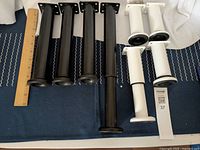 Both sets of metal extendable legs shown side by side including a ruler for scale, displaying four black and four white legs with mounting plates