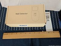 Plain brown box with 'Wall Detector' text and image of the stud finder device placed beside a wooden ruler showing approximate size.