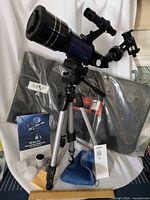 Full setup showing telescope mounted on silver and black tripod, instruction manual, lenses, color filters, cleaning cloth and carry case in background.