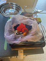 Three stainless steel mixing bowls nested together, clear glass measuring cup with wooden spoon, plastic bag containing various baking utensils, wood cutting board, black electric hot plate, metal baking tray, large glass baking dish with wire rack.