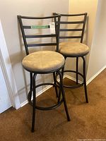 Two black metal bar stools with beige cushioned seats, positioned side by side against a wall, showing overall structure and style.